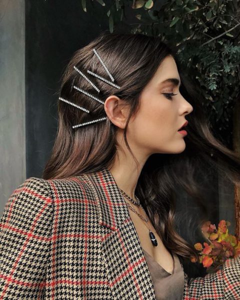 Mulher com cabelo castanho escuro e tranças decoradas com pentes de strass, usando blazer xadrez e acessório de colar, capturada em perfil com fundo natural e iluminação suave.