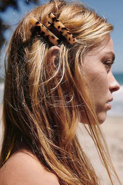 Mulher com cabelo castanho claro usando penteado com duas presilhas de cabelo de tigre, ao ar livre em um ambiente de praia.